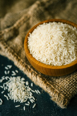 Close-up of raw white rice in a wooden bowl placed on a piece of burlap. Rustic background with scattered rice grains adds a natural and organic feel