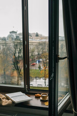 Person sitting by a window reading a Bible with a cup of coffee and a croissant on the windowsill, overlooking a rainy autumn park