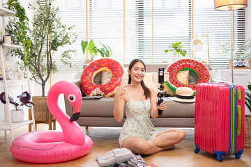 Young Asian woman  holding a camera and preparing for travel in a cozy living room, with a sofa, rubber ring, luggage, and sunglasses, summer time concept.
