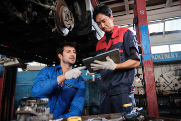 Two male mechanics cooperate, Latin and Asian workers fixing automotive wheels and maintaining car suspensions in garage station, and technician engineering service jobs in vehicle transport industry.