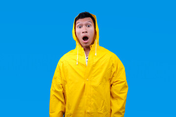 Shocked and surprised Asian man in waterproof raincoat with open mouth isolated on blue background © Gatot