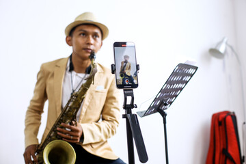 Young Asian Guy Influencer Play Saxophone Record With Smartphone For Online Audience Listen At Home