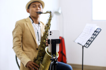 Fototapeta premium Selective Focus Of Smartphone Standing On Tripod For Recording With Young Musician Guy Playing Saxophone