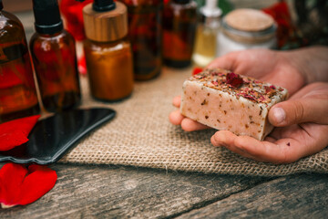 Close-up of hands holding a handmade floral soap bar topped with dried rose petals, with blurred amber essential oil bottles on a burlap-covered wooden surface