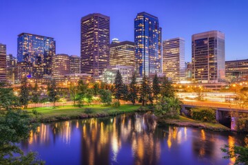 Fototapeta premium City skyline at twilight, reflecting in a river with parkland Modern high-rises, illuminated at night, mirrored in calm waters City park and roadway are visible