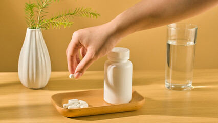 A hand holds a white pill and lifts it off a tray, underneath are many other pills and an unlabeled pill bottle placed on a wooden tray. A clear glass filled with clean water. Brown background