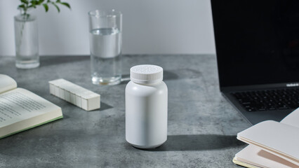Mockup of a medicine bottle with no label and placed on a gray desk. On the desk are a laptop, a notebook, a book and a medicine box. A functional food medicine bottle with a therapeutic effect.