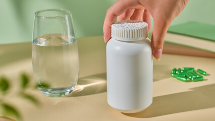 Front view of an unlabeled pill bottle being lifted off a table by a hand. Several soft green pills are placed in the background. Foreground has blurred foliage. Natural light falls on objects.