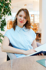 Portrait of a confident businesswoman sitting at a table in a modern office or coworking space. She is smiling warmly and looking at the camera while holding an open notebook. 