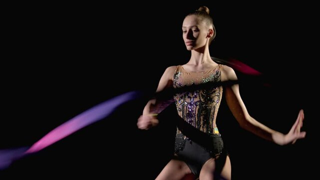 Portrait of graceful attractive rhythmic gymnast in elegant leotards with patterned ribbon performing snakes elements on black background.