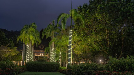 Tropical resort park in the evening. Rows of royal palm trees along the alley. The trunks are wrapped with decorative night lighting. The hotel building is far away. Green vegetation. Dark sky.