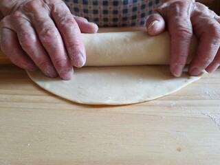 An elderly lady rolls out dough with a wooden rolling pin on a table. Close-up of the elderly lady's hands. Home cooking
