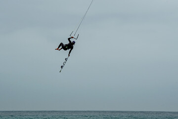 Fototapeta premium Kite surfers in the waves on a windy day. Tropical beach huts and spectators watching on the sandy beach, Bright colourful kites fill the cloudy sky. Turquoise water. copy space. Sport vacation.