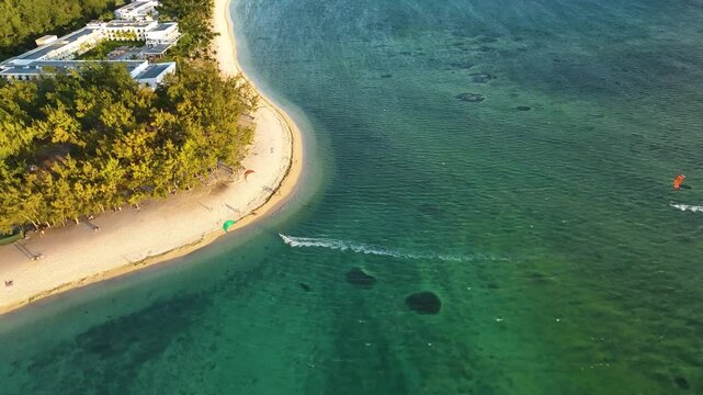 Aerial view of two kitesurfers gliding along shallow green-blue waters near a luxury beach resort surrounded by trees in Mauritius