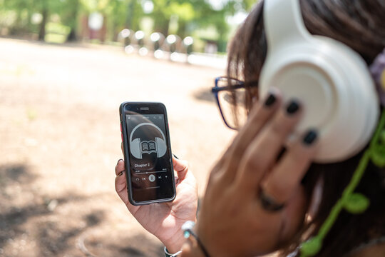 Woman listening to audiobook, relaxing on park bench with wireless headphones and smartphone during sunny day