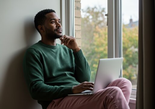 Man with laptop sitting by window looking thoughtful indoors