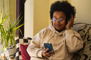African American woman wearing headphones and listening to music via smartphone while sitting on...