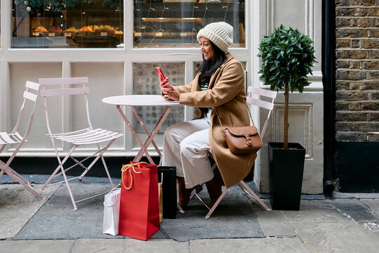 Woman sitting at an outdoor cafe using a smartphone with shopping bags during Christmas.