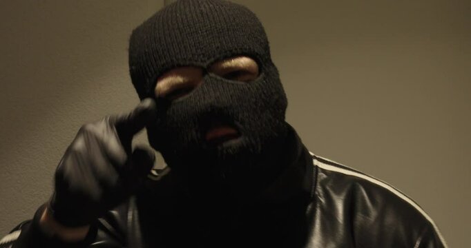 A man wearing a mask and black gloves speaks to the camera, pointing his finger. He threatens and shouts.