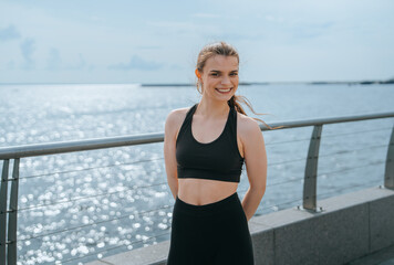 Fototapeta premium Smiling woman poses confidently in sports attire by a glistening ocean under a bright sky.