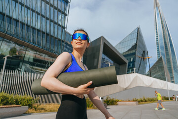 A confident woman strides with a yoga mat under her arm, wearing sunglasses, against a backdrop of futuristic skyscrapers.