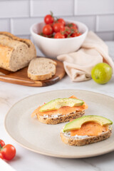 Two whole grain toasts with cream cheese, salmon and avocado served on a beige plate, with bread, cherry tomatoes and a lime in the background. Healthy breakfast in a minimalist style