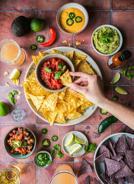 Top view hand dipping chip in salsa with guacamole, drinks on tile.