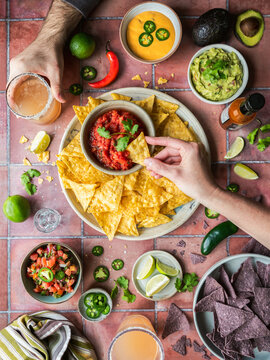 Top view hands having chips, salsa, guacamole, drinks on tile.