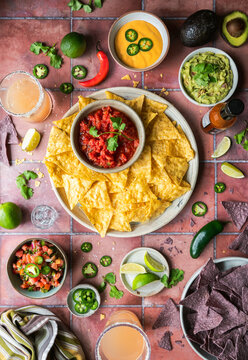 Top view tortilla chips, salsa, guacamole, queso and drinks on tile.