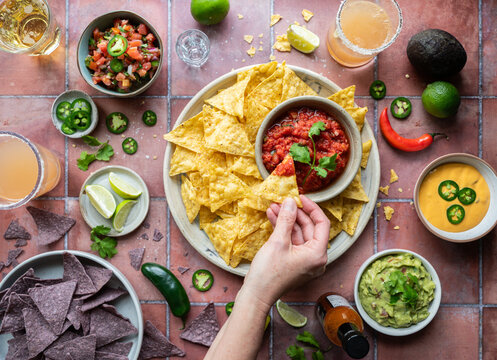 Top view hand dipping chip in salsa with guacamole, drinks on tile.