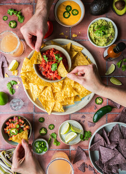 Top view hands with chips, salsa, guacamole, drinks on tile.