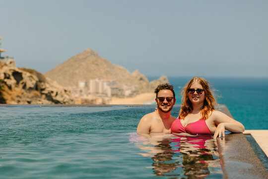 Couple leaning on infinity pool edge, smiling with ocean backdrop