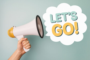 Hand holding megaphone announcing "Let's Go!" in a speech bubble, against a pale-green background, showcasing motivation and announcement concepts