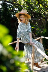 Beautiful girl in dress and hat sitting on wooden bridge in summer in garden