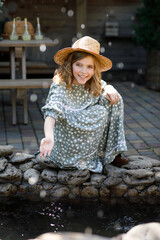 Beautiful happy girl in dress and hat sits in garden by pond and plays with water in summer