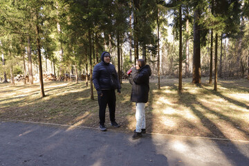 A man and woman pose on a forest trail in warm winter clothing, with the sun casting soft shadows across the leaf-strewn path