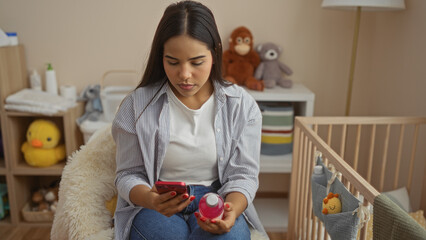 Woman using phone holding baby bottle in cozy nursery with baby items around in soft-lit room,...