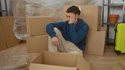Young man sitting among cardboard boxes in a new home shows a thoughtful expression in a sparsely...