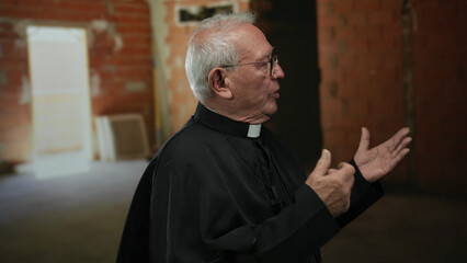 Senior priest in black robe gestures thoughtfully indoors at a brick-walled construction site,...