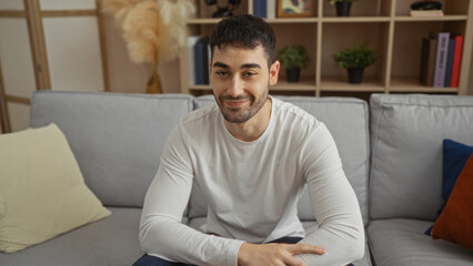 Fototapeta premium Young man sitting indoors on a sofa in a cozy living room with plants and books, looking relaxed and handsome in a home environment.