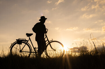 Silhouette of a man farmer wearing a hat, holding a bicycle in his hands at sunset