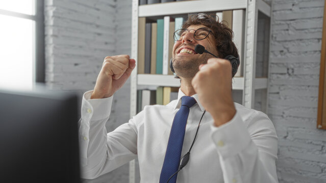 Young man celebrating success in office with headset and shirt, expressing happiness in modern workplace by bookshelf, showing winning gesture energetically.