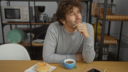 Young man sitting in an office, thoughtfully gazing while enjoying coffee and a donut, with modern interior visible in background, implying a relaxed workplace atmosphere.