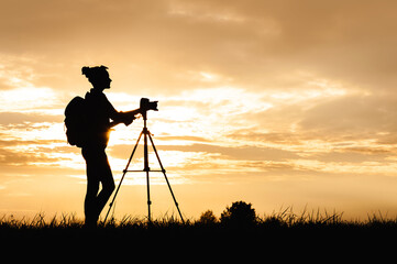 Silhouette of a female photographer with a photo-video camera on a tripod, standing in a meadow in a field. Background of sunset on the sky.