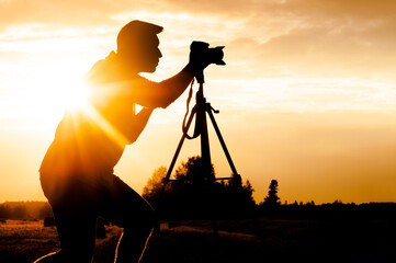 Silhouette of a photo-video camera standing on a tripod, with a lifestyle male photographer filming...