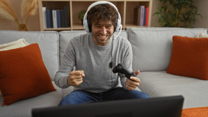 Young man gaming in cozy living room with headphones and controller, expressing excitement on a comfortable sofa, surrounded by decor and books