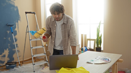 Young man in a new home using a laptop while painting the interior walls, suggesting a modern diy...