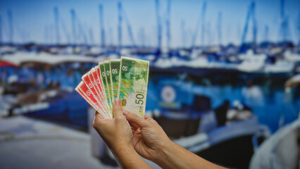 Man holding israeli shekel banknotes on a seaside dock with boats visible, depicting a financial investment concept in an outdoor coastal setting in israel.