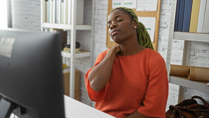 Woman in orange shirt with braids sits relaxed in modern office touching neck amidst bookshelves and documents, creating a serene professional ambiance indoors.