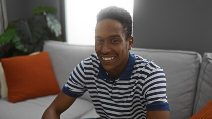 Young man sitting in a living room, smiling warmly, wearing a striped shirt and surrounded by cozy home decor, creating an inviting and relaxed atmosphere indoors.
